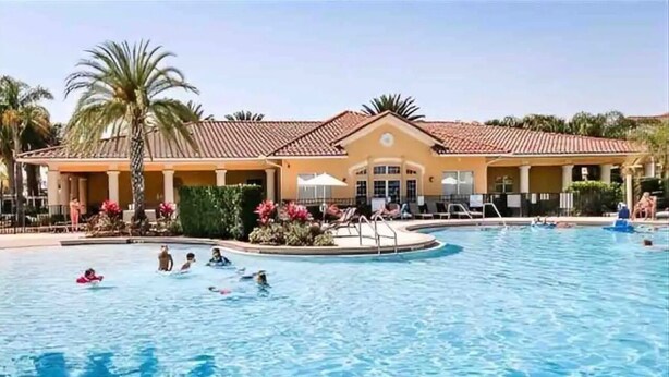 A swimming pool with people, a palm tree, and a building with a tiled roof. - Amazing Disney Themed Oasis 3br Near Parks 3 Bedroom Townhouse by RedAwning (Kissimmee)