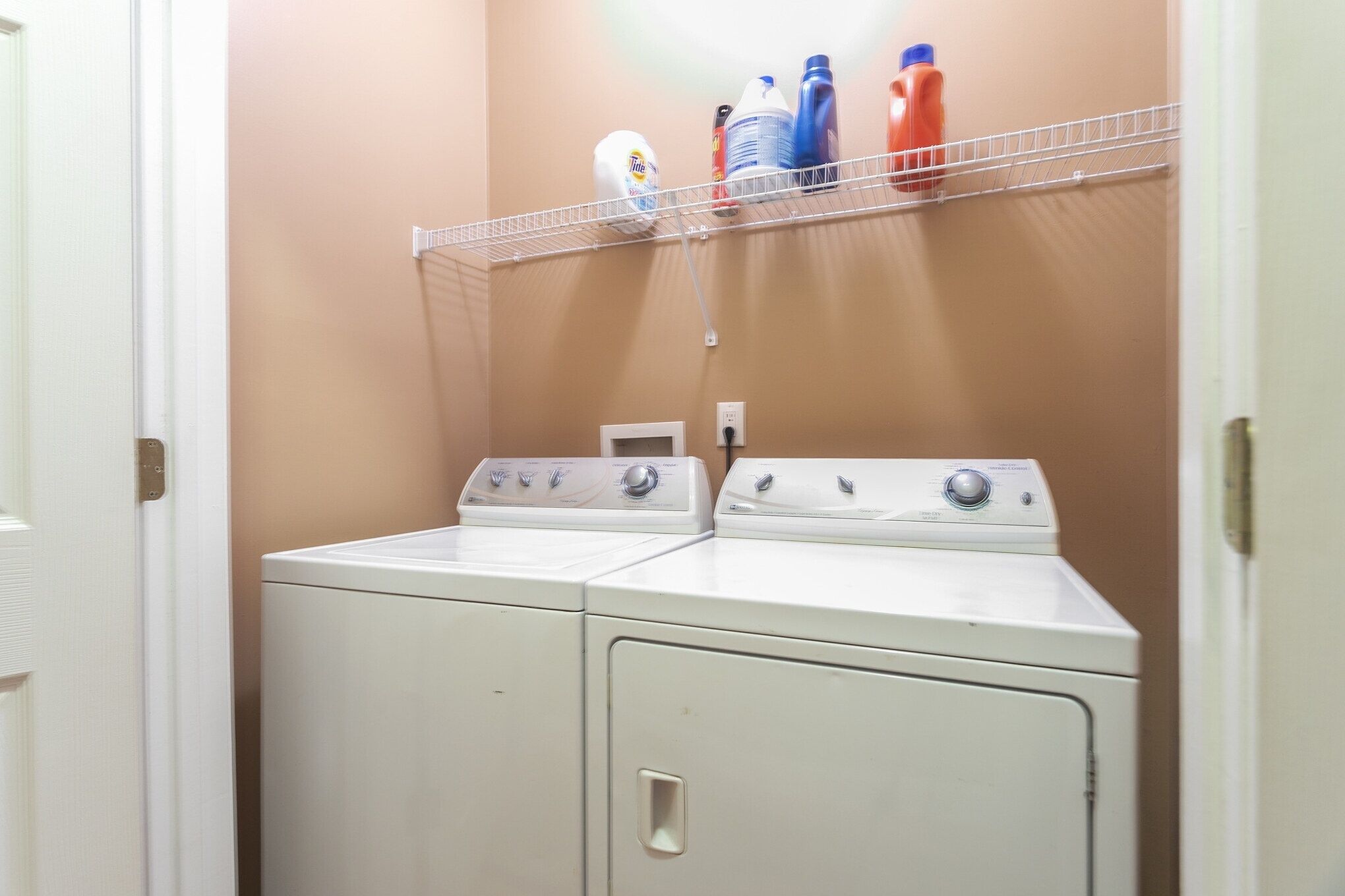 A laundry area with two stacked washers and dryers, a metal shelf above holding detergent bottles, and a white door on the left.