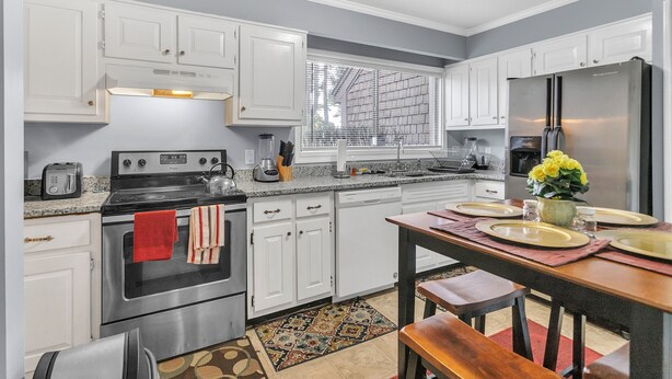 A modern kitchen with white cabinets, stainless steel appliances, and a dining area with a wooden table and bench. - Centrally Located 4br Stonecrest Condo 4 Bedroom Condo by RedAwning (Lithonia)