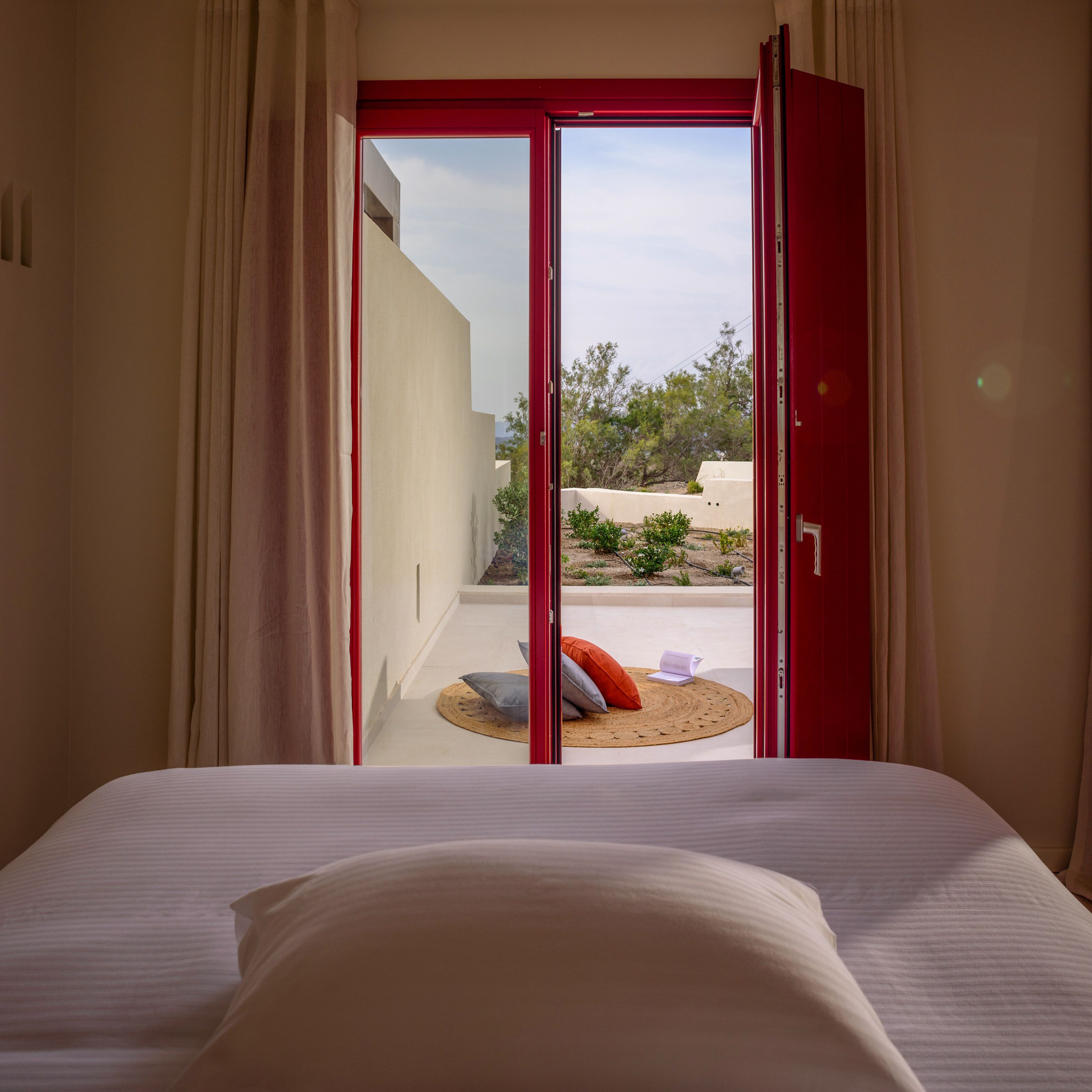 Un dormitorio con cama, vista a través de una puerta de marco rojo hacia un espacio exterior con una pequeña mesa y sillas.