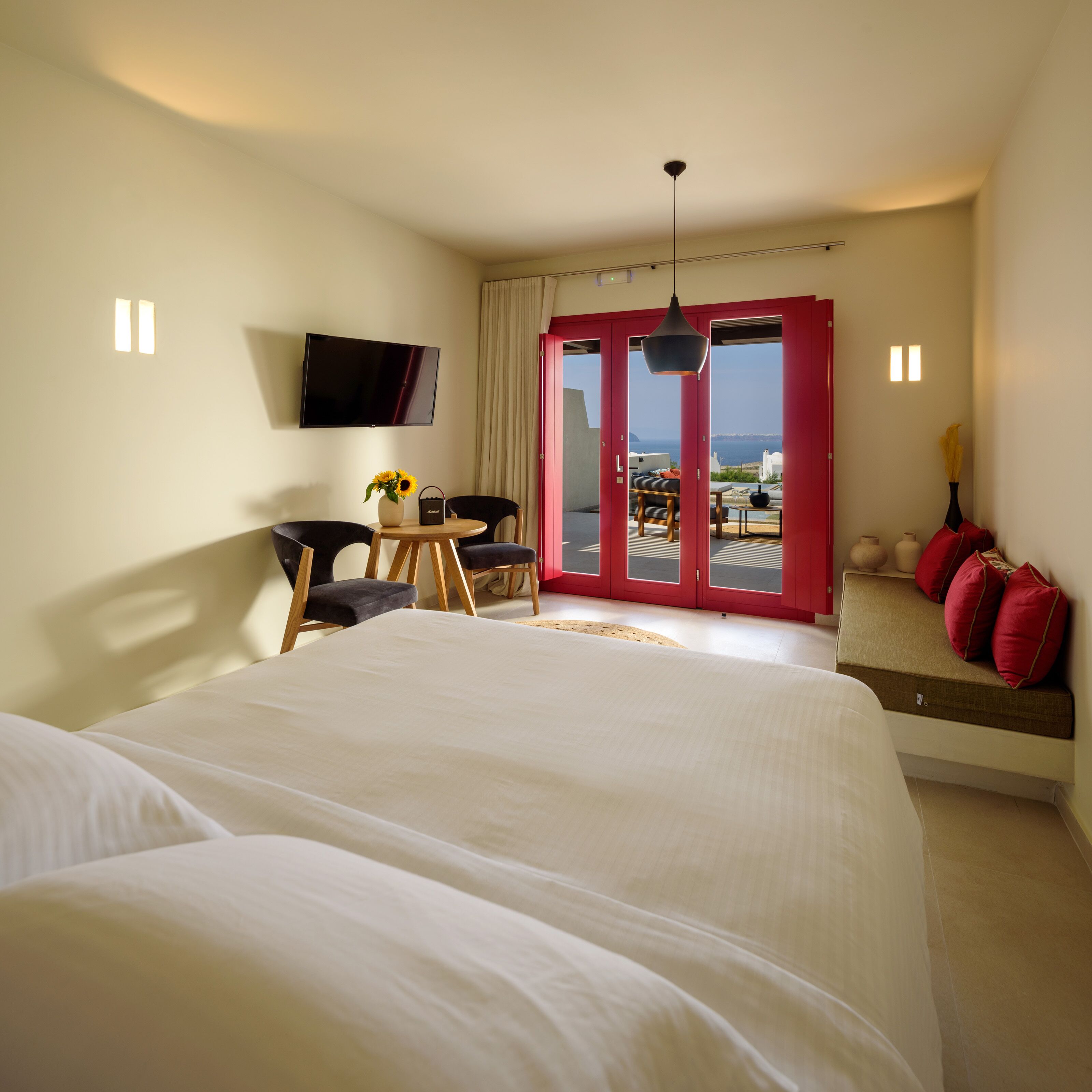 A hotel room with a large bed, a desk with a chair, a TV mounted on the wall, and a view of the sea through a glass door.