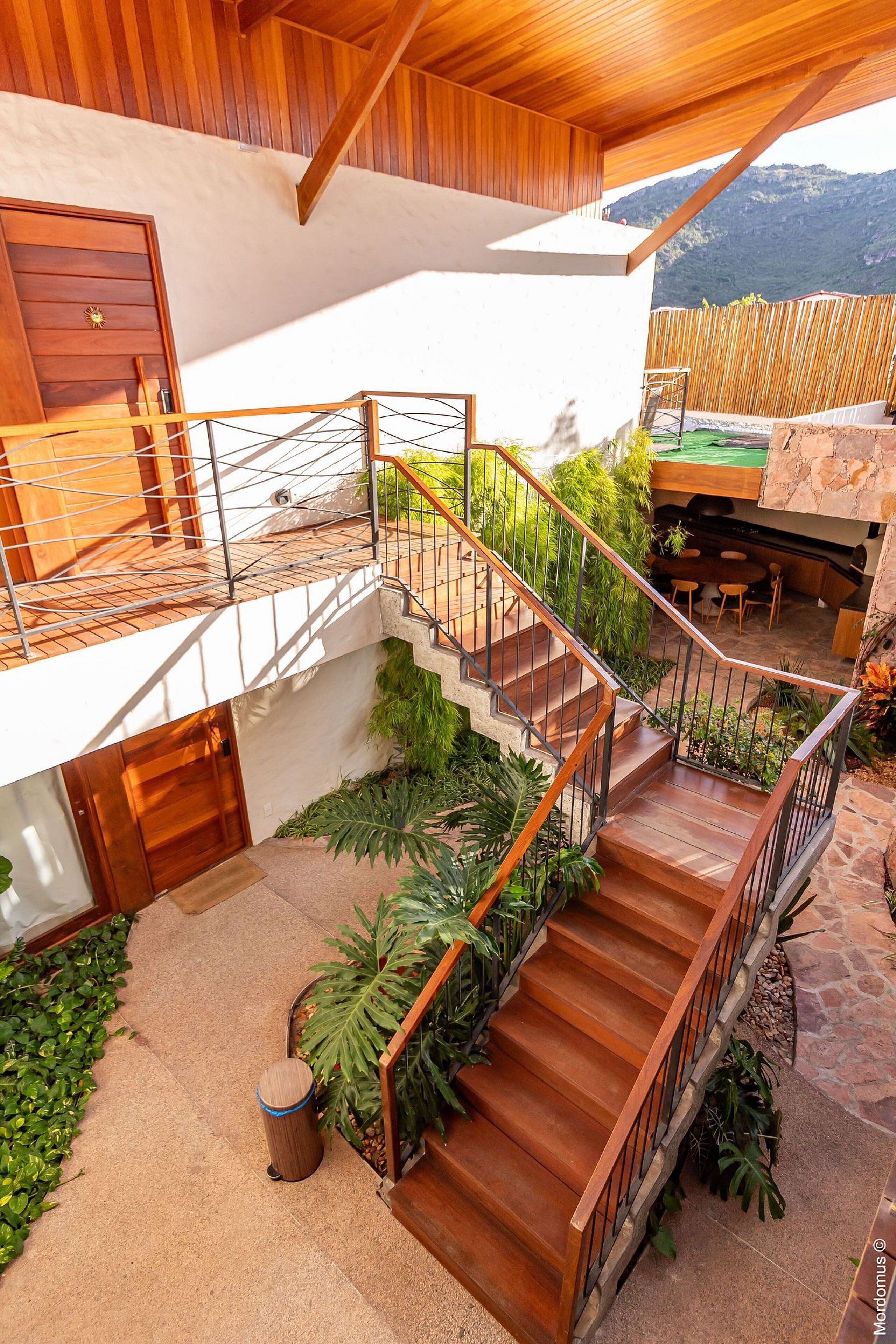 Uma casa moderna com escada de madeira que dá para uma varanda, rodeada por vegetação e uma parede de pedra.