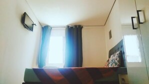 Una habitación pequeña y escasamente amoblada, con una cama individual, una ventana con cortinas y un televisor montado en la pared.