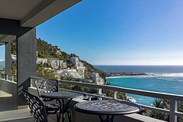 A balcony with two tables and chairs overlooking a coastal view with buildings on a hillside.