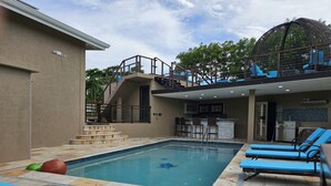 Un área de piscina con una piscina cristalina, sillas de descanso azules y un bar con taburetes.