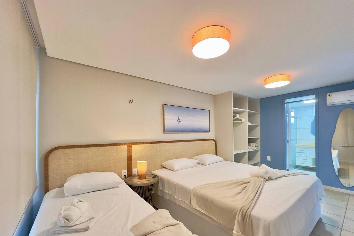 A hotel room with two beds, a bedside table with a lamp, a wardrobe, and a bathroom with a mirror.