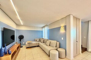 Uma sala de estar moderna com um sofá seccional, uma televisão de tela plana, um ventilador de pedestal e um console de madeira.