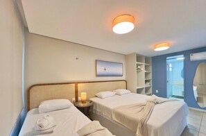 Quarto de hotel com duas camas, uma mesa de cabeceira com luminária, guarda-roupa e banheiro visível através de uma porta aberta.