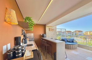 A modern kitchen with a bar area, a large window with a view of buildings, and a balcony with a wicker sofa.