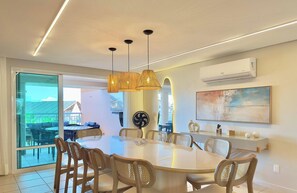 Uma área de refeições com mesa de vidro, cadeiras de vime, iluminação pendente e um ar condicionado na parede.
