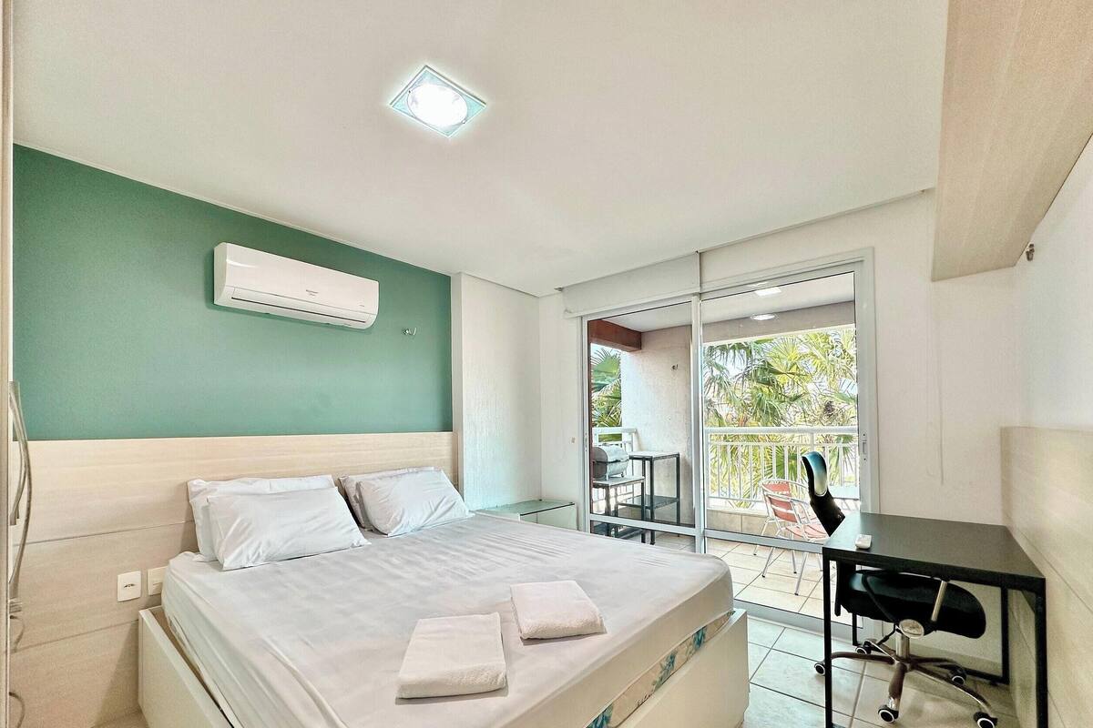 A hotel room with a bed, a desk, a chair, a balcony with a table and chairs, and an air conditioning unit.