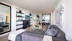A modern living room with a grey sofa, a glass coffee table, and a fireplace. There is a kitchen area visible in the background with a dining table and chairs. - YOLO Spaces Clifton - Clifton Beachfront Magic Apartment (Cape Town)