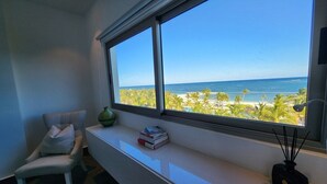 A room with a large window overlooking a beach, a white desk, a chair, and a small table with books and a vase. - Condominio Aquamarina (Punta Cana)