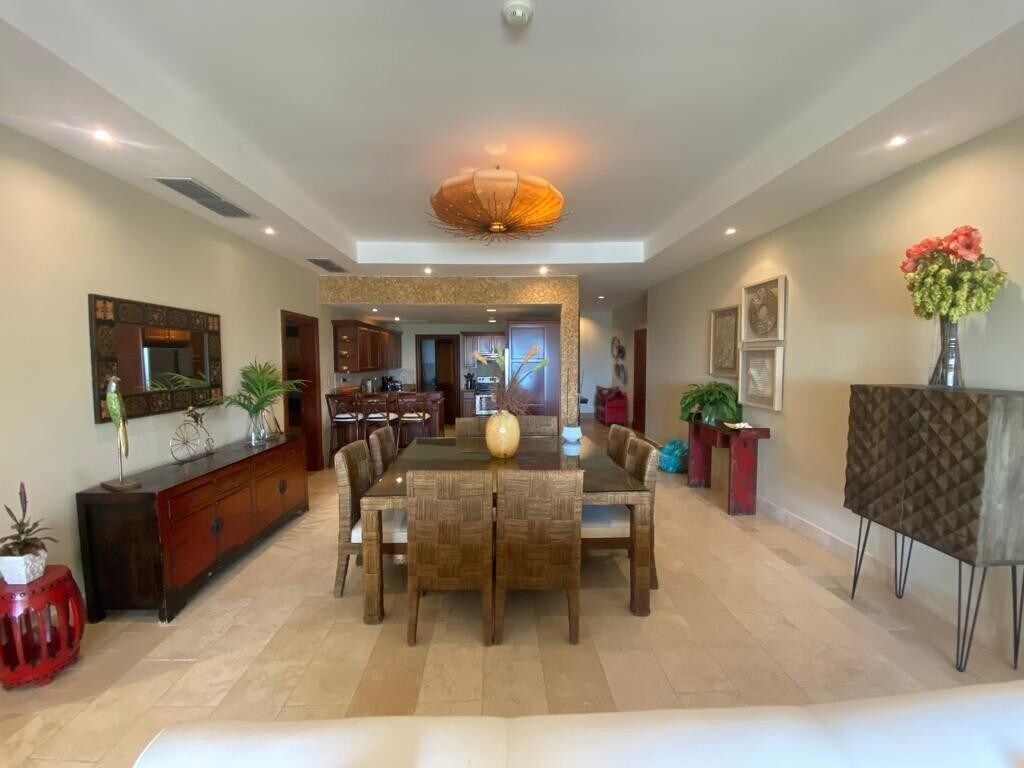 A spacious dining area with a large wooden table and chairs, a decorative ceiling light, and a view into the kitchen.