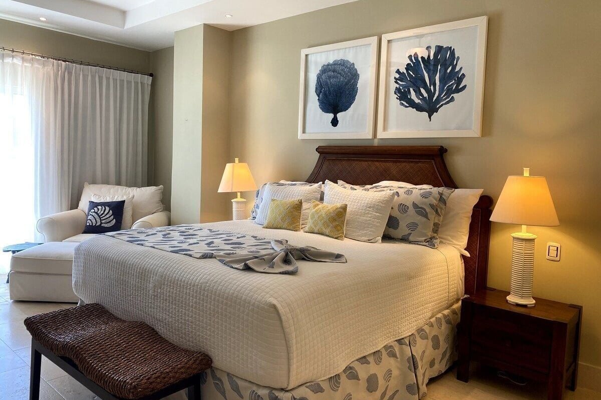 A hotel room with a large bed, two bedside lamps, a chair, a bench, and two framed wall art pieces.