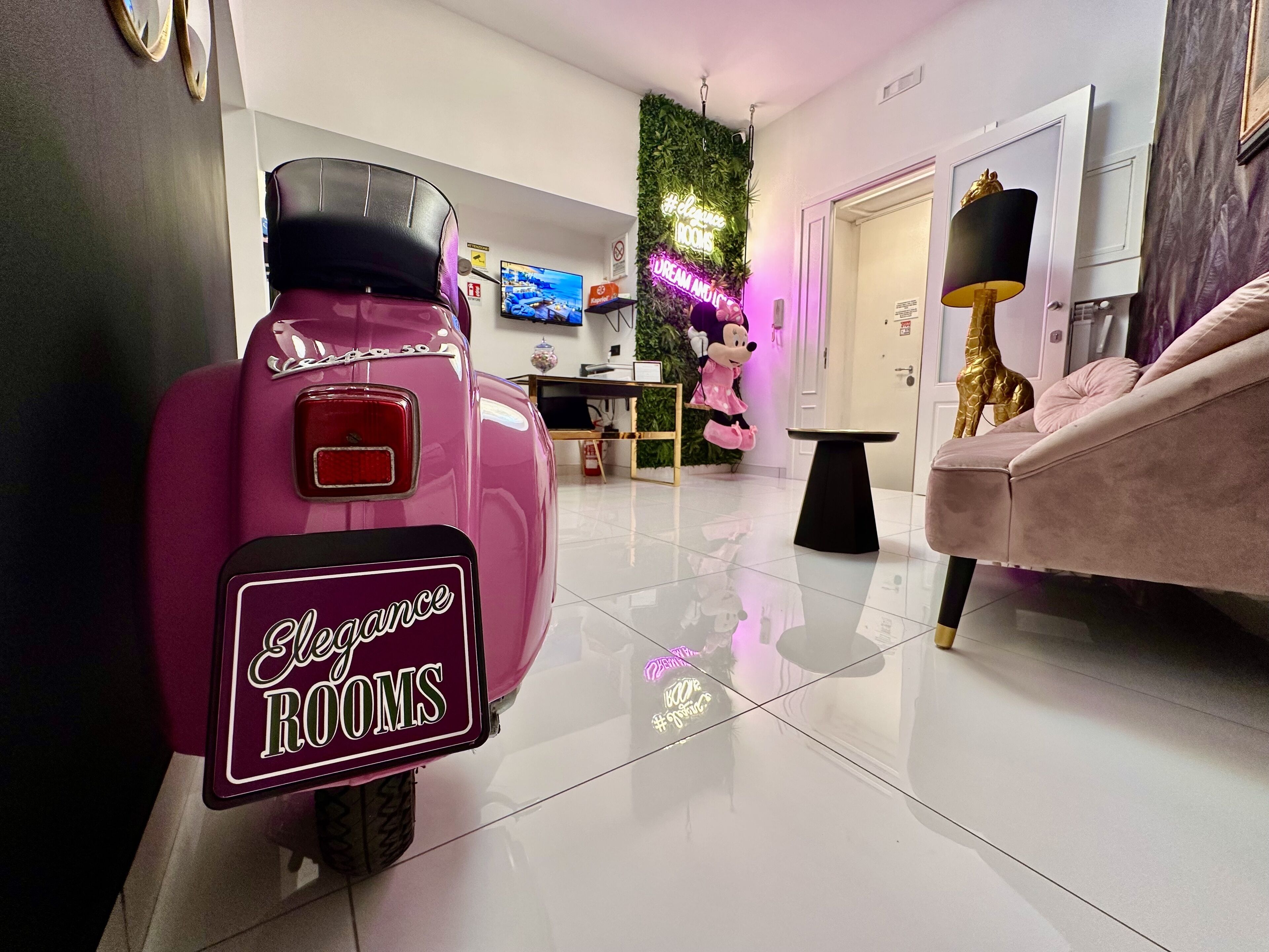 A pink scooter with a neon sign reading "Elegance Rooms" parked in a modern, well-lit room with a white floor, a plush sofa, and a small table.