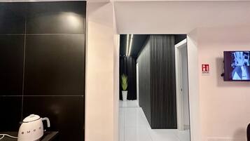 Ein moderner Hotelgang mit Wand in schwarzer Fliesenoptik, weiß gefliestem Boden, einem Blumentopf, einem Brandschutznak und einer Überwachungskamera.