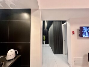 Un couloir d’hôtel moderne avec un mur recouvert de carreaux noirs, un sol carrelé de blanc, une plante en pot, un panneau d’alarme incendie et une caméra de surveillance.