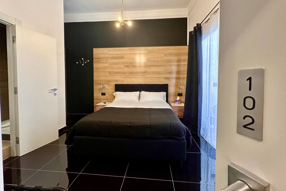 Une chambre d’hôtel moderne avec un grand lit, une tête de lit en bois, une table de chevet, une lampe et une fenêtre avec des rideaux.