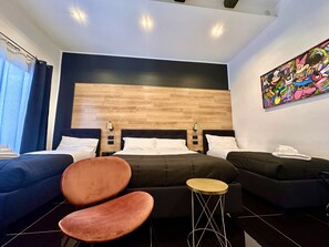 Une chambre d’hôtel moderne avec un grand lit, deux tables de chevet, un canapé, un fauteuil et un tableau coloré accroché au mur.