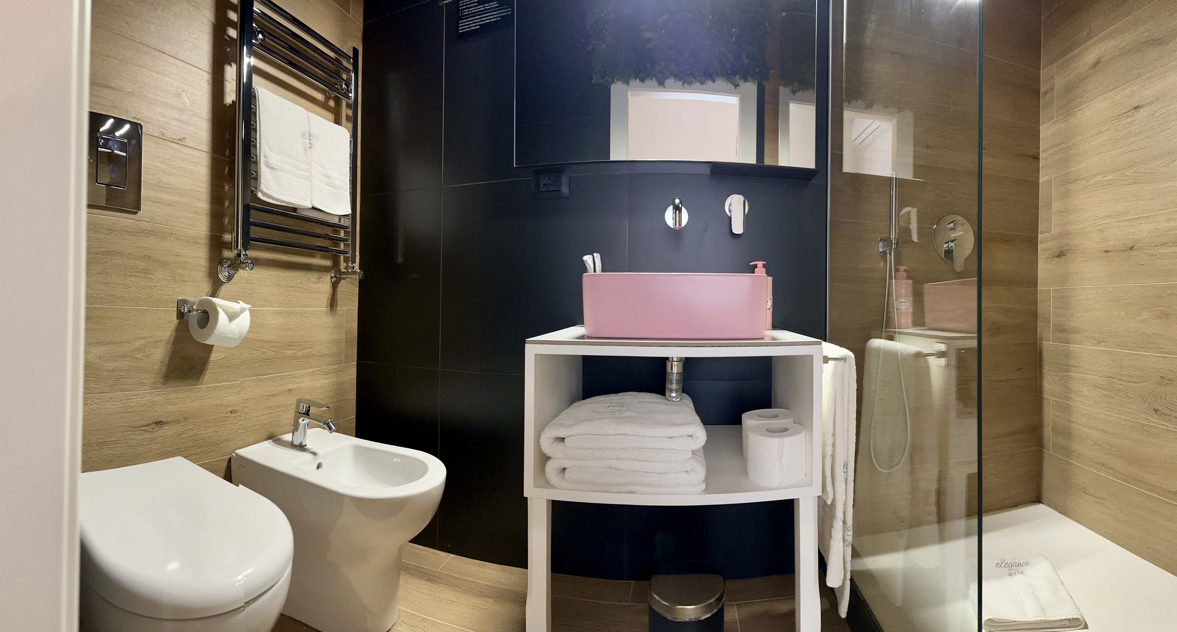 Un baño moderno con un bidé, un lavamanos con dispensador de jabón rosa, una estantería con toallas y una mampara de cristal para la ducha.