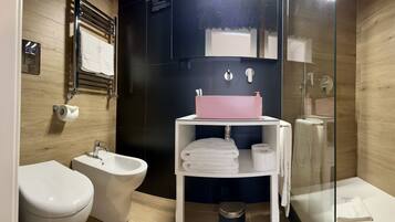 Un baño moderno con un bidé, un lavamanos con dispensador de jabón rosa, una estantería con toallas y una mampara de cristal para la ducha.