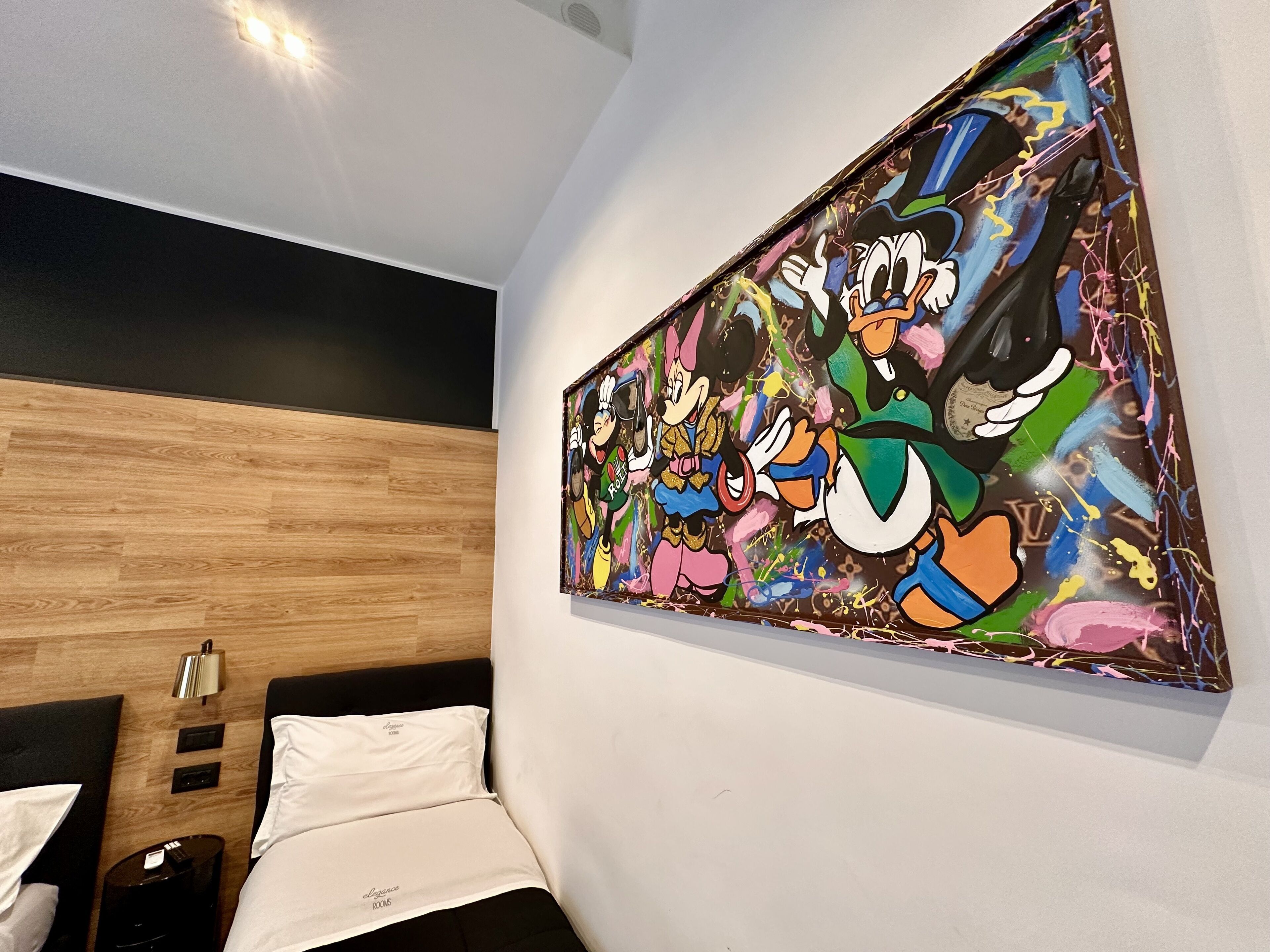 Une chambre d’hôtel avec une tête de lit en bois, un lit recouvert de draps blancs, une table de chevet avec une lampe et une grande fresque colorée sur le mur.