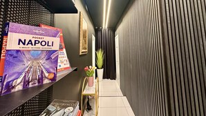 A narrow hallway with a shelf displaying travel brochures, a potted plant, and a framed picture on the wall. - Elegance Rooms Spaccanapoli (Naples)