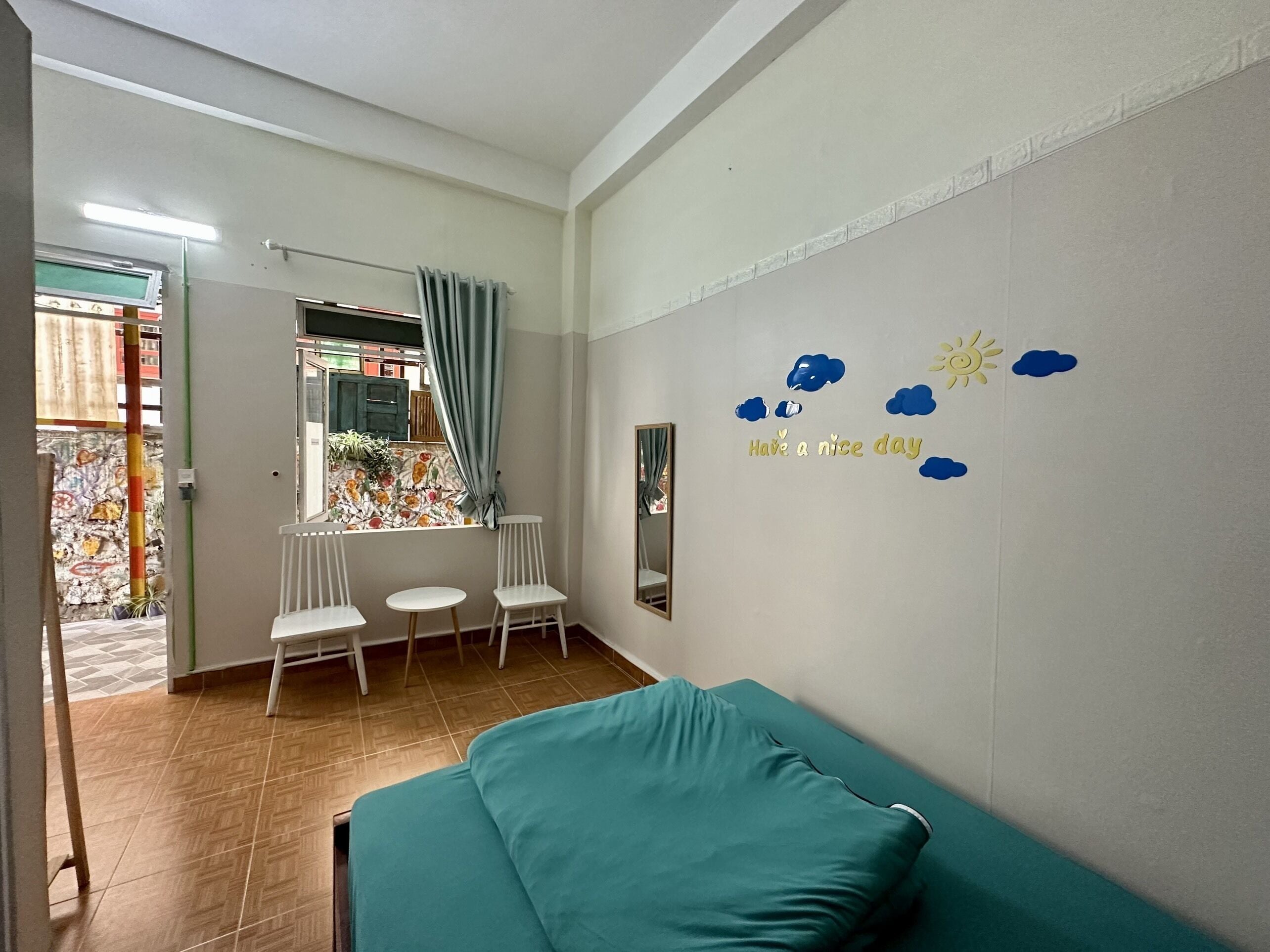 A room with a teal bed, white chairs, a small table, a window with curtains, and a wall with a 'Have a nice day' message.
