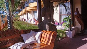 A wooden bench with pillows, a hammock, and a large tree sculpture in a courtyard with a building in the background. - Hotel Kima (Bombinhas)