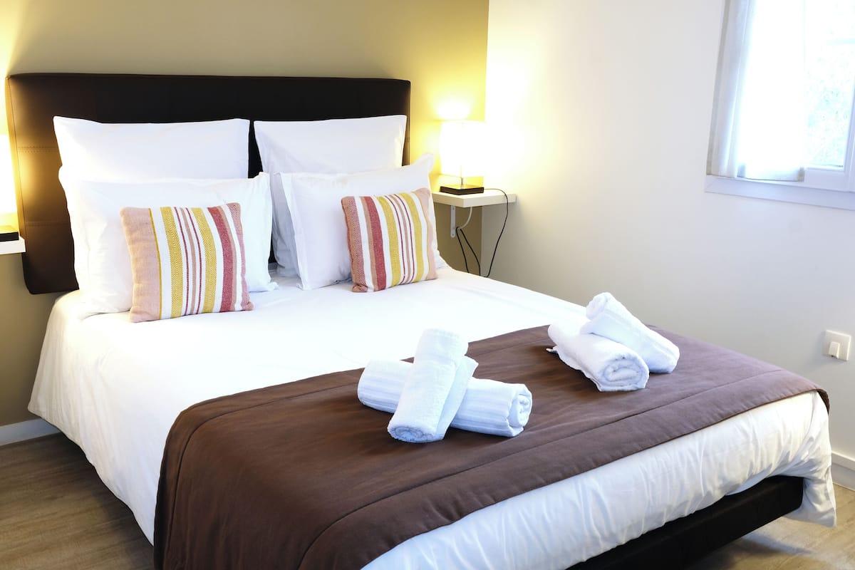 A neatly made bed with white and striped pillows, a brown bedspread, and rolled towels on the side.