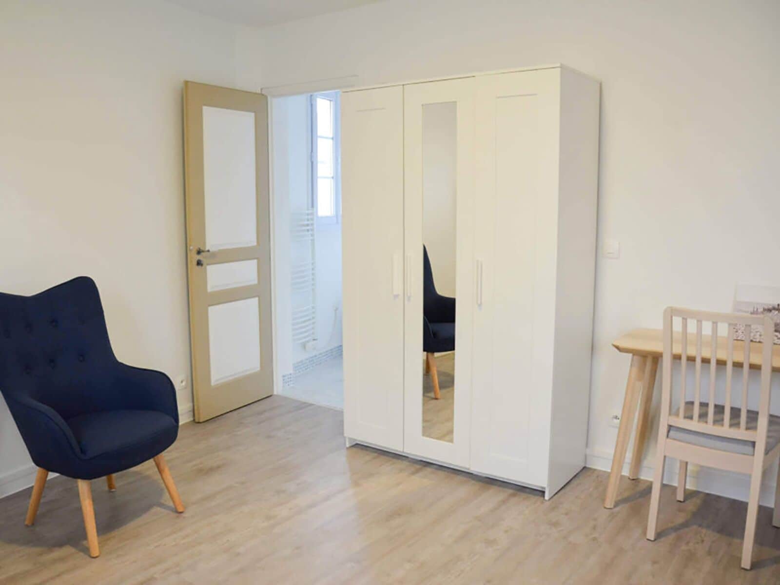 Een kamer met een kledingkast, een donkerblauwe stoel, een houten tafel en een witte deur.