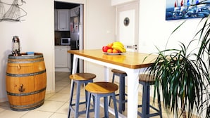 Une cuisine avec une table en bois, des tabourets, un panier de fruits, un tonneau et une plante en pot.