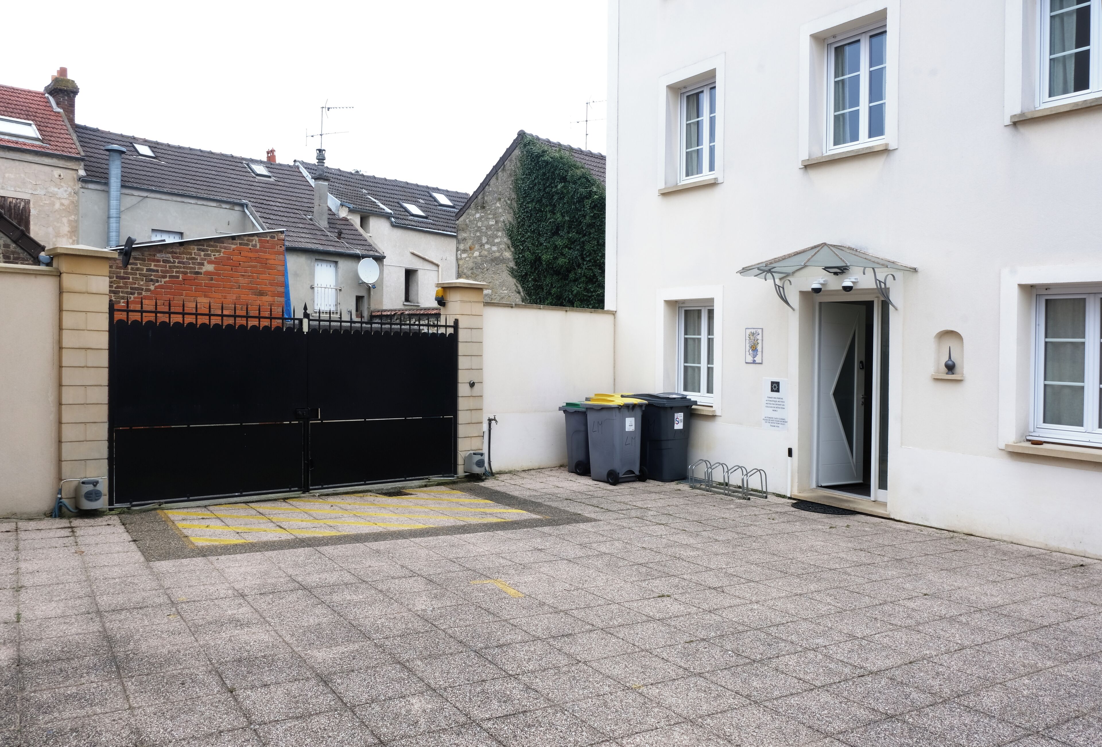 Une cour fermée par un portail noir, un bâtiment blanc avec une porte, et deux poubelles.