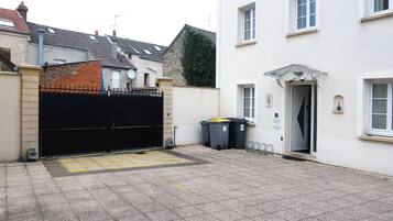 Une cour fermée par un portail noir, un bâtiment blanc avec une porte, et deux poubelles.