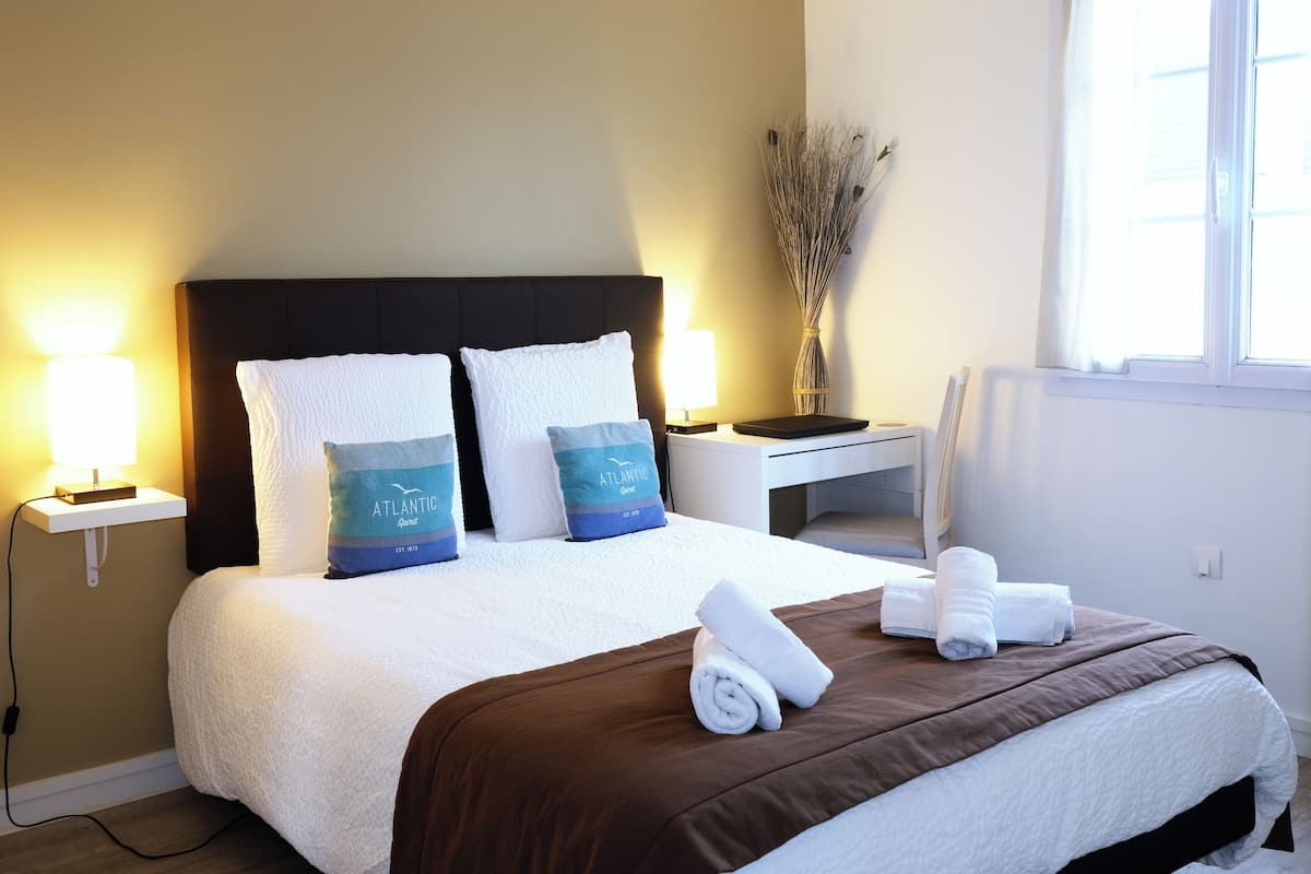 A neatly made bed with white and blue pillows, a brown bedspread, and rolled towels on the side.