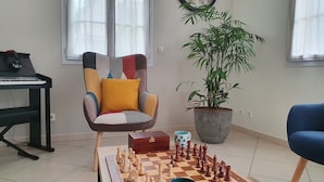 Un salon avec un jeu d’échecs, un piano, un fauteuil, une plante et une horloge au mur.