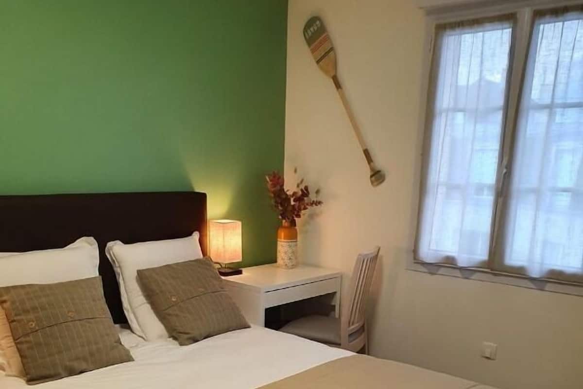 A neatly made bed with a beige blanket and pillows, a bedside table with a lamp and vase, and a window with curtains.