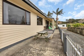A house with a covered patio, a wooden picnic table, and a fenced garden area. - Seaside on Stratford - Cable Bay Holiday Home (Cable Bay)