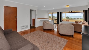 Ein modernes Wohnzimmer mit einem braunen Sofa, beigen Sesseln und einem großen Teppich. Holzdielenboden, eine Tür und ein Blick ins Freie durch große Fenster.