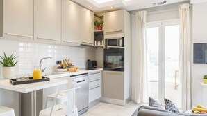 A modern kitchen with white cabinets, a built-in oven, and a microwave. There is a dining area with a white chair and a small table. - Dimi Brou's Apartments by CorfuEscapes (Corfu)