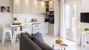 A modern kitchen with white cabinets, a dining area with white chairs, a grey sofa, and a small table with fruits and a book. - Dimi Brou's Apartments by CorfuEscapes (Corfu)