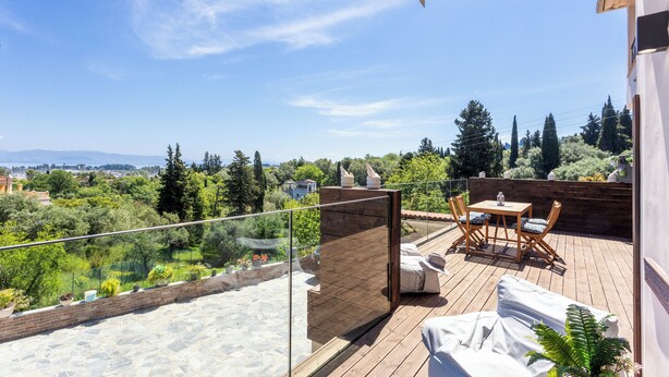 A wooden deck with a glass balustrade, outdoor furniture, and a view of greenery and distant buildings. - Dimi Brou's Apartments by CorfuEscapes (Corfu)