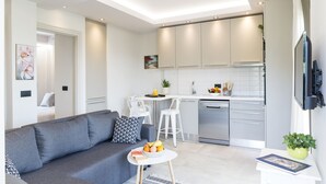 A modern living room with a grey sofa, a small white table with fruit, and a kitchen area with white cabinets and a dishwasher. - Dimi Brou's Apartments by CorfuEscapes (Corfu)