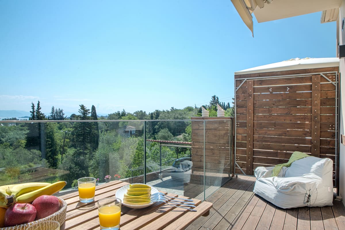 A balcony with a wooden table, fruit basket, and drinks, overlooking a forested area.