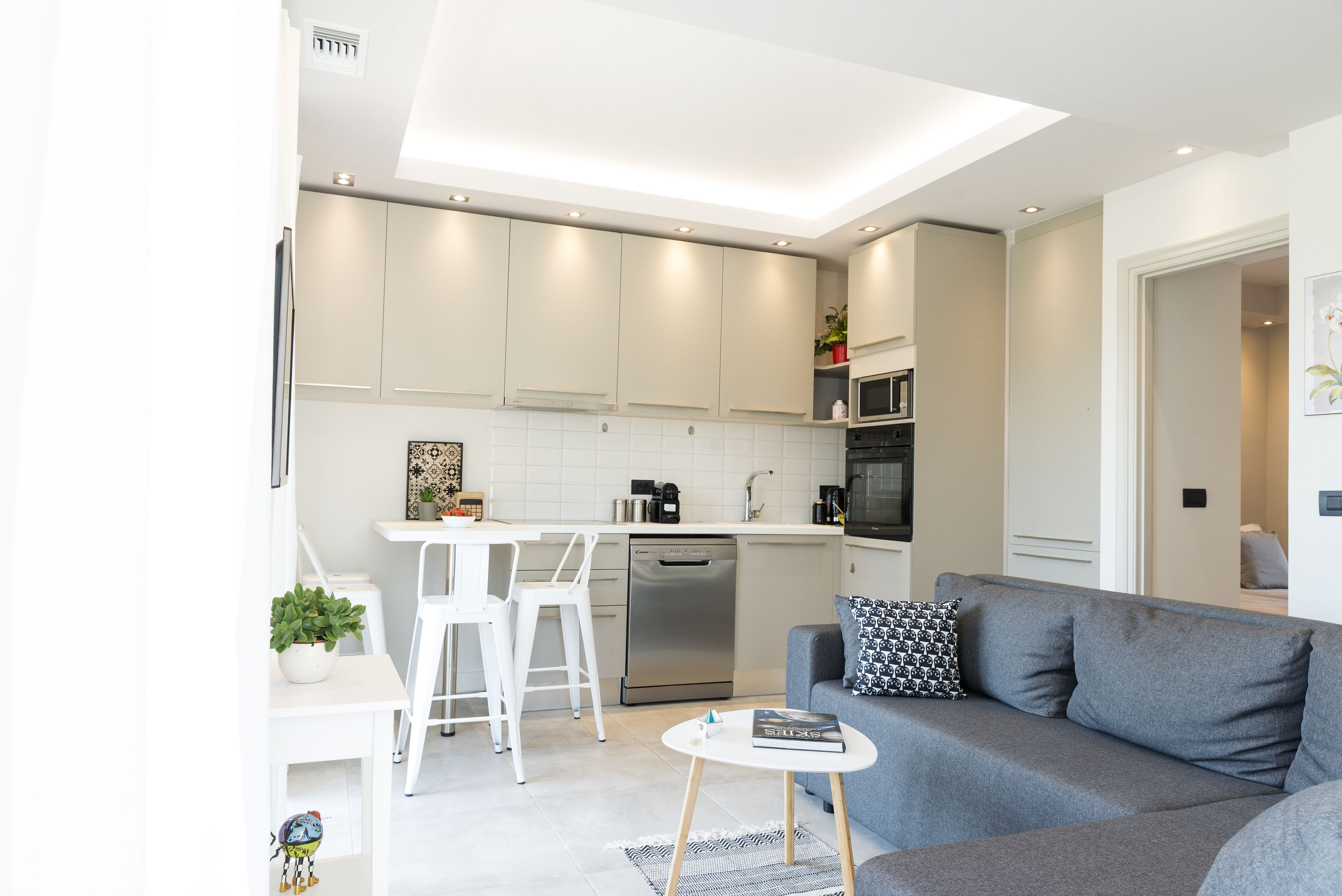A modern kitchen with white cabinets, a built-in microwave, and a dishwasher. There is a grey sofa with a black and white pillow, a small white table, and a plant on the counter.