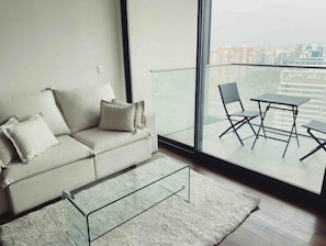 Uma sala de estar moderna com um sofá, uma mesa de centro de vidro e vista para um horizonte urbano através de grandes janelas.