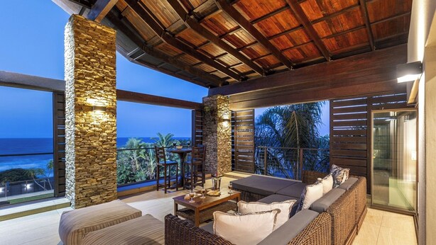 A modern outdoor seating area with stone walls, a wooden ceiling, and a view of the ocean.
