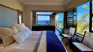 A bedroom with a large bed, a view of the ocean, and a balcony with a chair.