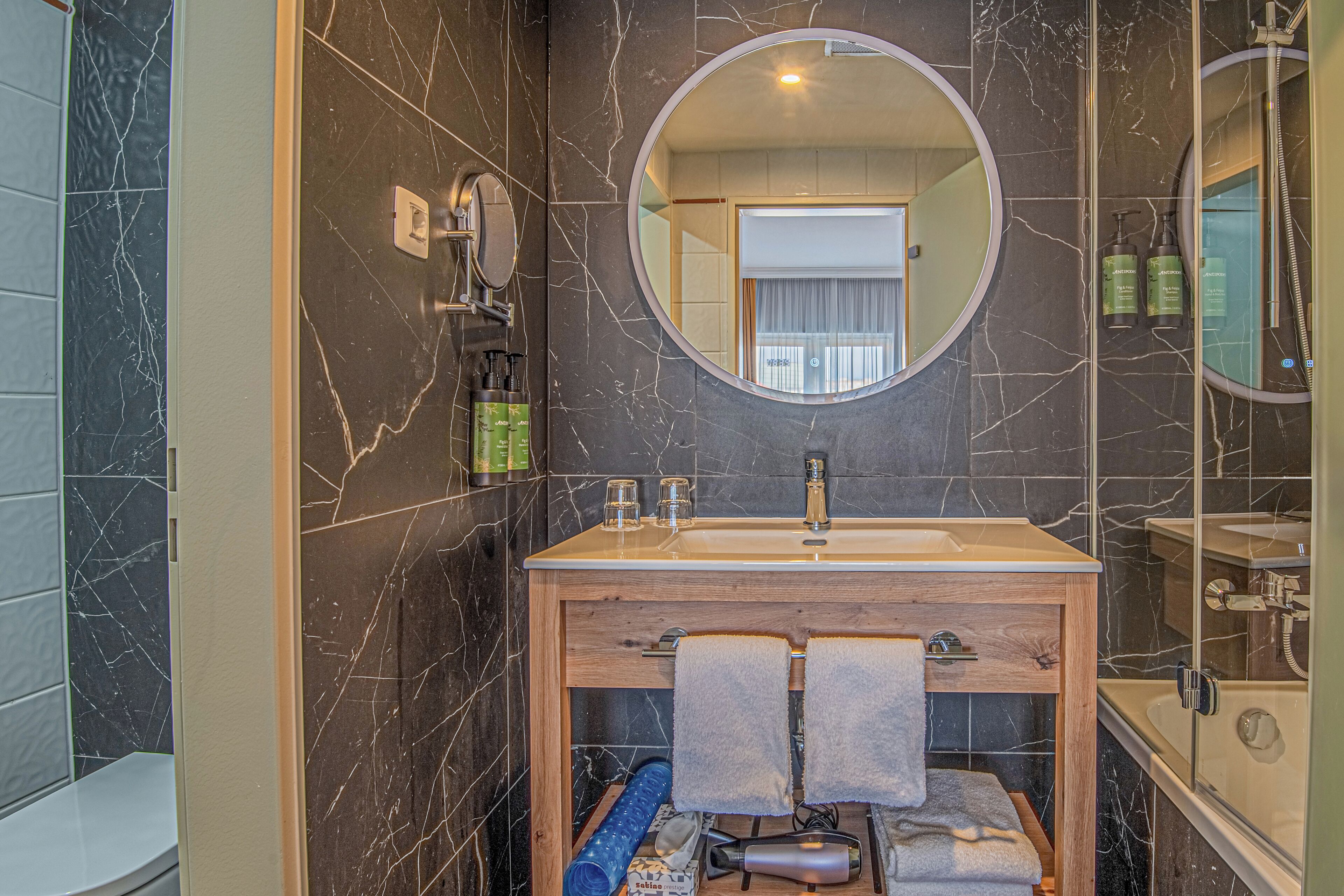 Un bagno con una parete in marmo, un mobile lavabo in legno, uno specchio rotondo e una vasca da bagno.